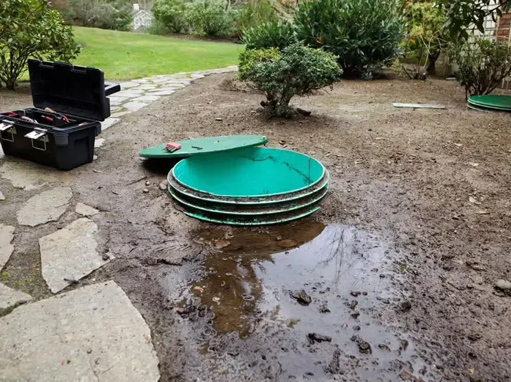 An open septic tank lid with wastewater pooled on soaked ground beside a house